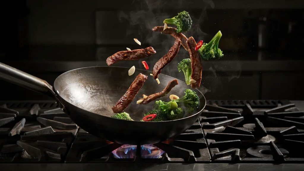 Steaming pieces of seared steak and broccoli florets tossed in the air above a cast iron wok on a gas stovetop.