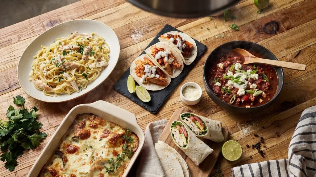 Top-down view of chicken pasta, tacos, baked casserole, and wraps served on a wooden dinner table.