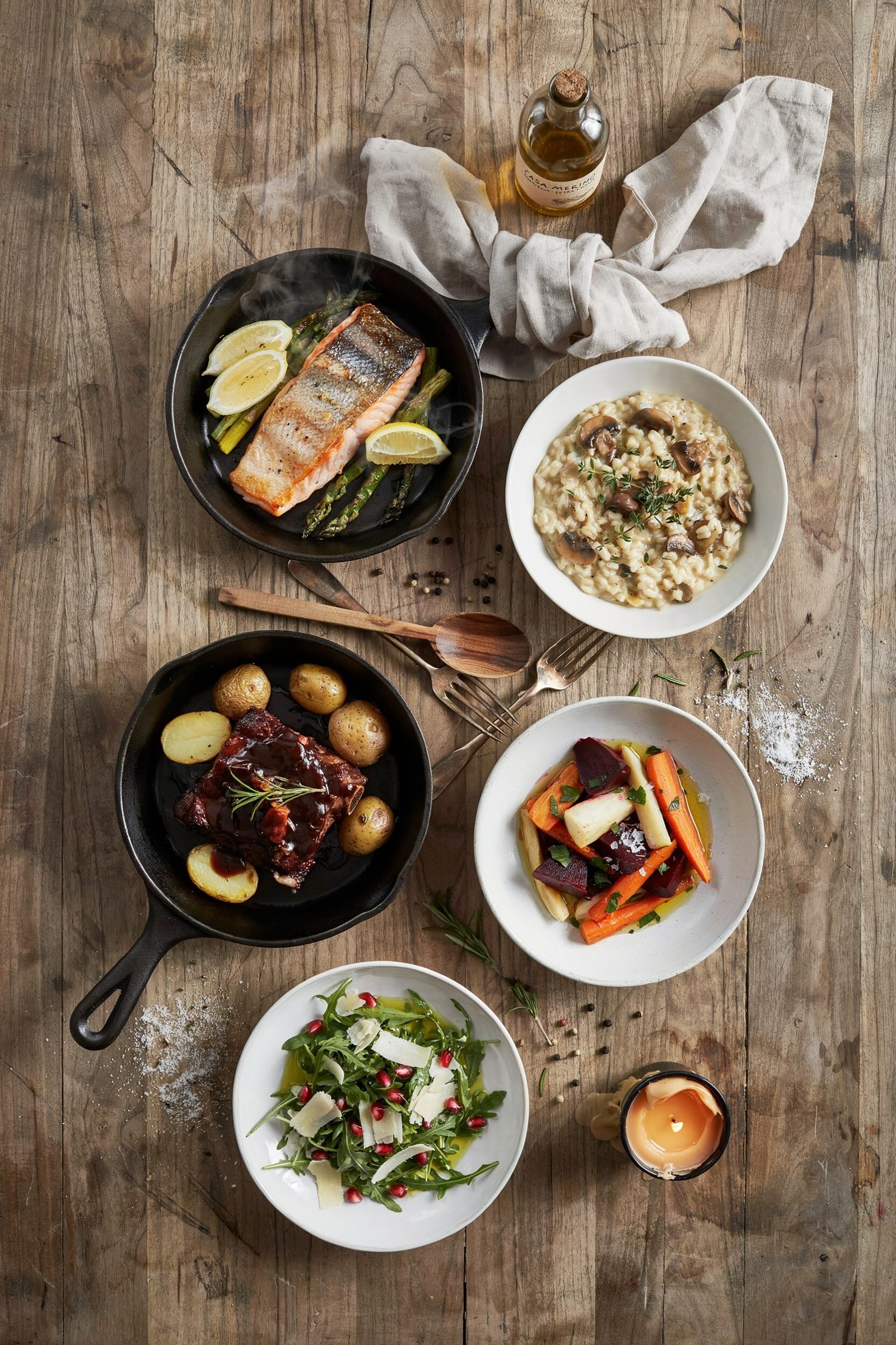 Top down view of a wooden table set with salmon, risotto, ribs, root vegetables, and a salad.