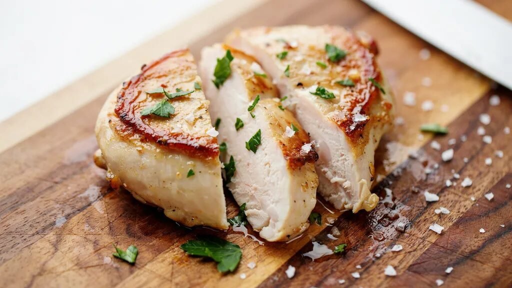 Close-up of a tender sliced chicken breast topped with fresh herbs and sea salt flakes on a wood board.