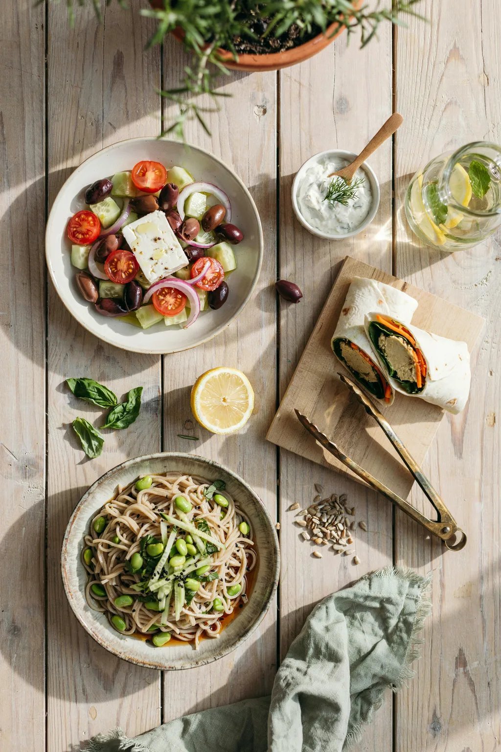 Top view of a Greek salad, edamame noodles, and veggie wraps served on a rustic light-colored wooden table.