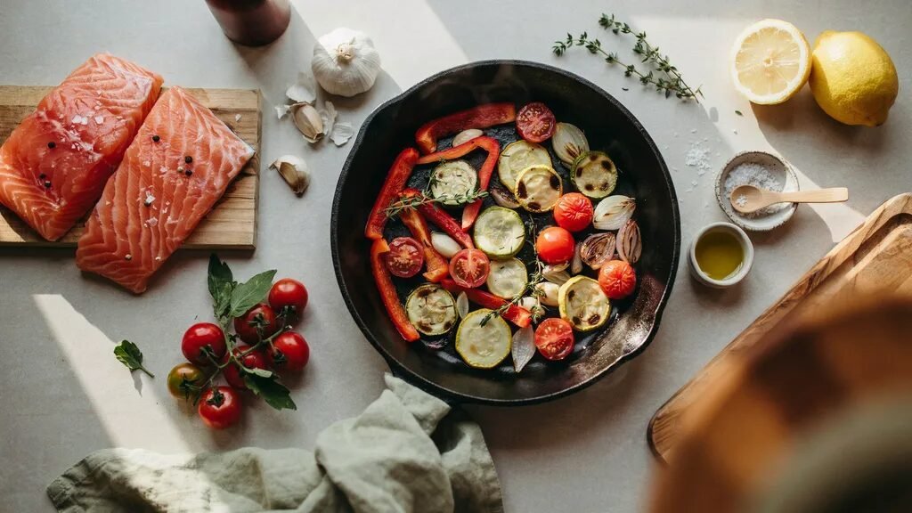 Cast iron skillet filled with roasted peppers, zucchini, and tomatoes next to raw salmon slices and lemon.